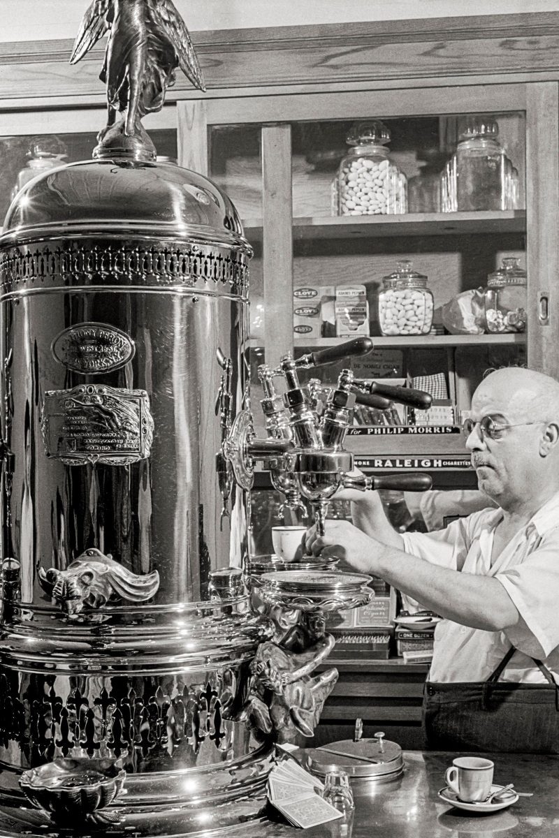 Muž pripravuje kávu na vintage espresso stroji v retro kaviarni s dreveným interiérom a potravinovými nádobami.
