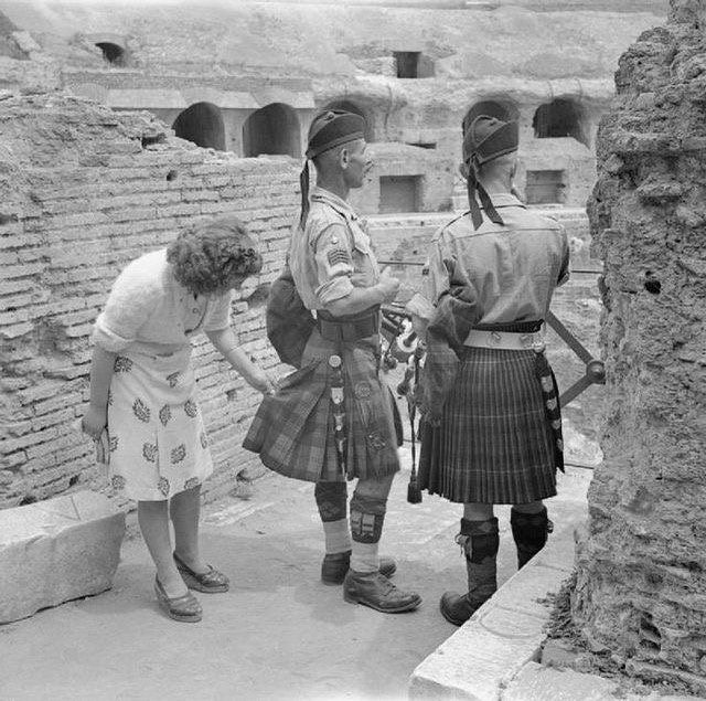 Žena skúma tradičné škótske kilty dvoch vojakov v historickej lokalite z kamenných blokov.