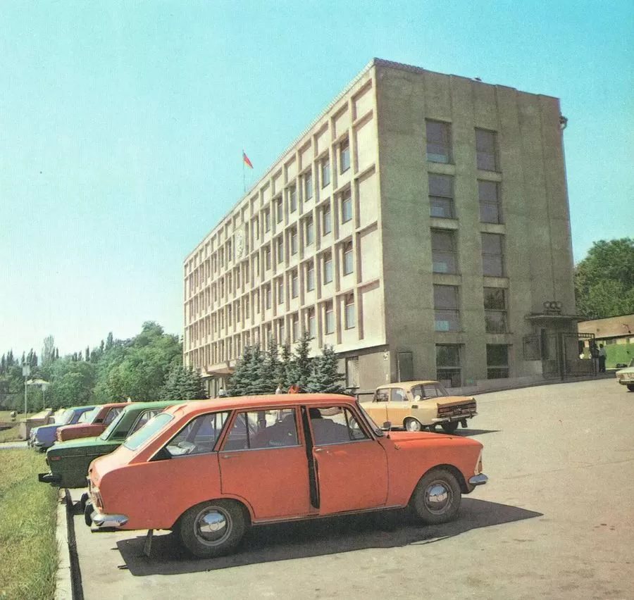 Parkovisko s autami pri veľkej administratívnej budove, modrá obloha, letný deň.