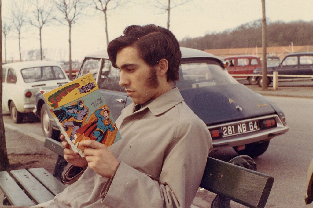 Osoba sedí na lavici a číta komiks Superman a Batman, okolo sú zaparkované retro autá.