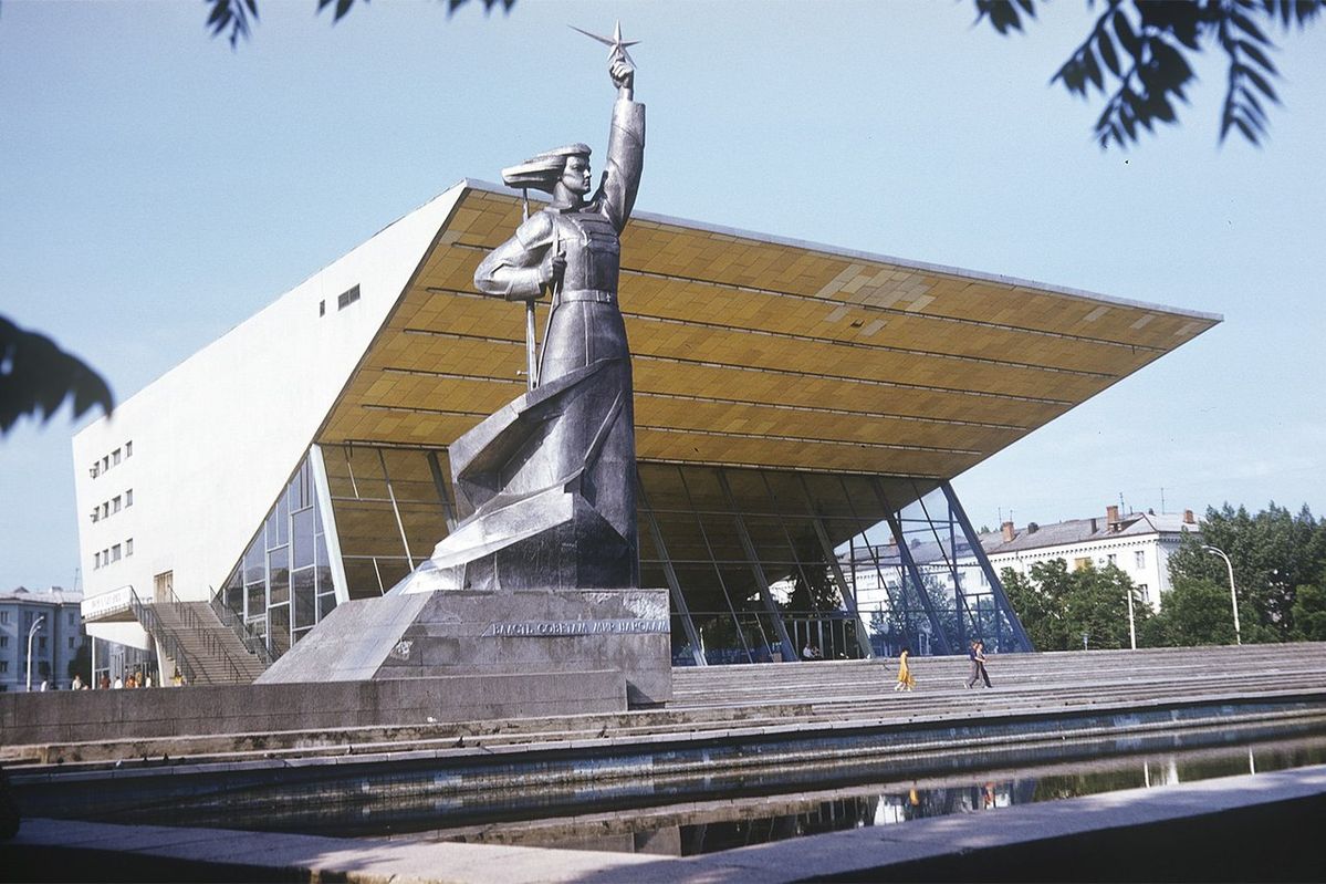 Moderná socha vojaka pred architektonicky výraznou budovou na námestí, modrá obloha v pozadí.