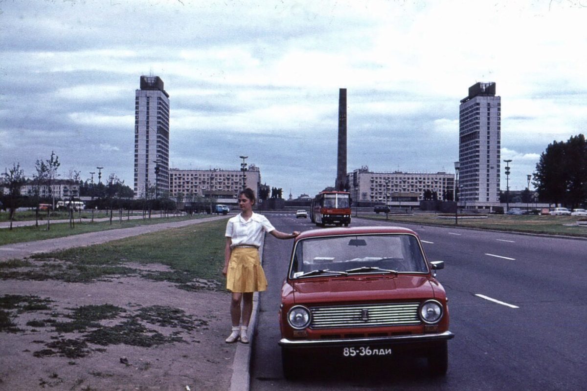 Žena v žltej sukni stojí pri červenom aute na širokej ceste v mestskom prostredí so socialistickou architektúrou.