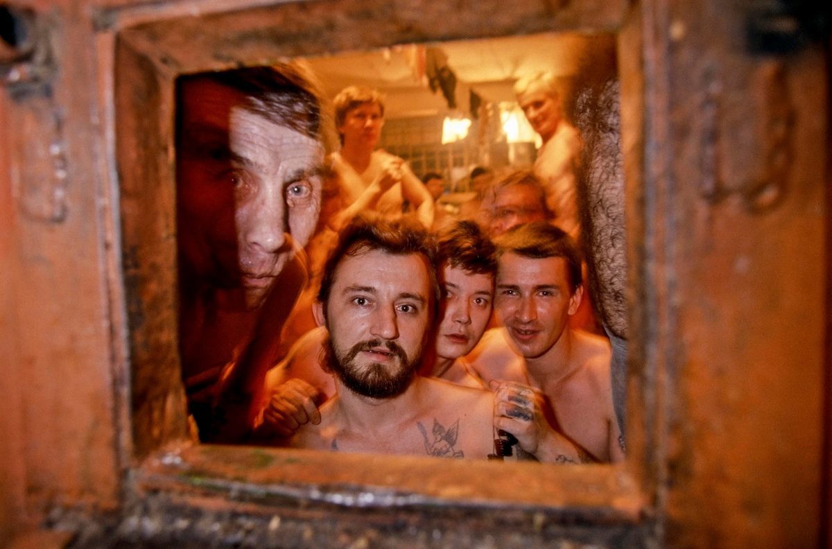 Skupina ľudí pozerá do objektívu cez otvor v stene, všetci v tesnom priestore, zdá sa, že sú v saune.