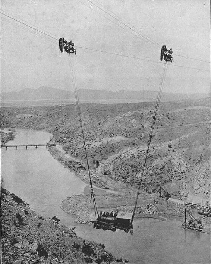 Vozík na lanách prepravuje pracujúcich ponad roklinu so širokou riekou v pozadí. Historická preprava cez kaňon.