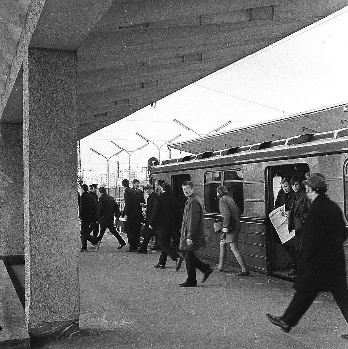 Cestujúci vystupujú z vlaku na nástupišti, čiernobiela fotografia zo železničnej stanice.