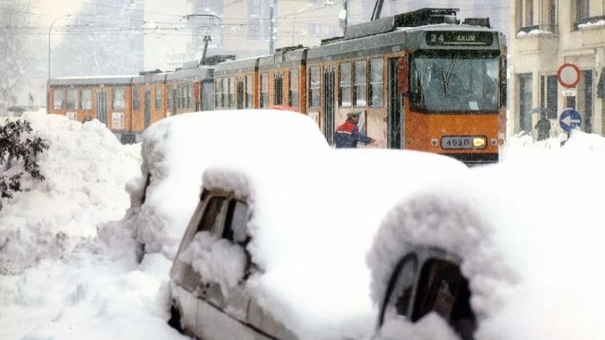 Električka prechádza zasneženou mestskou ulicou, autá pokryté snehom.