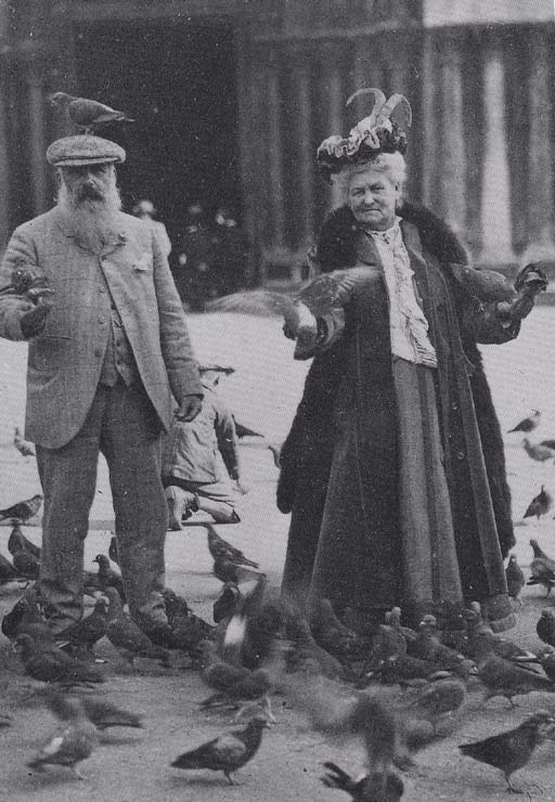 Starší pár kŕmi holuby na námestí, muž má holuba na klobúku. Vintage fotografia.