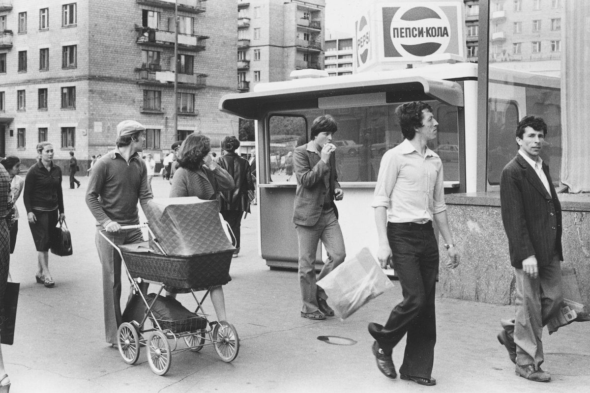 Skupina ľudí na ulici pred stánkom s logo Pepsi, historická mestská scéna s kočíkom.