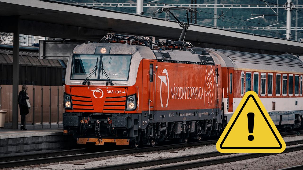Červený vlak na stanici s varovným symbolom, ktorý upozorňuje na dôležité oznámenie alebo zmenu v doprave.
