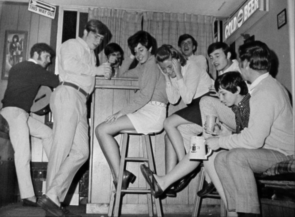 Skupina priateľov v 70. rokoch, zabávajúc sa pri bare s pivom a úsmevmi. Retro atmosféra a móda.