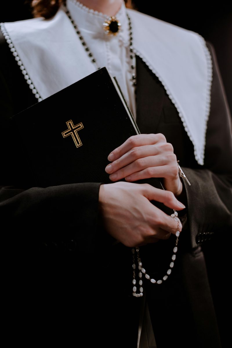 Osoba v obleku drží čiernu Bibliu so zlatým krížom a ruženec na perlovom náhrdelníku.