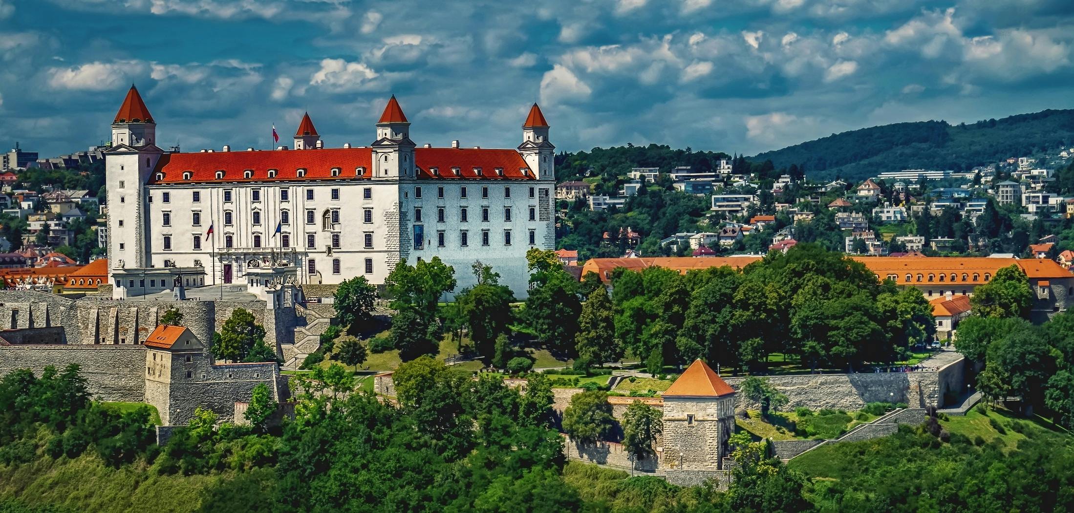 Hrad na kopci so štyrmi vežami, obklopený zeleňou a mestom na pozadí pod modrou oblohou.