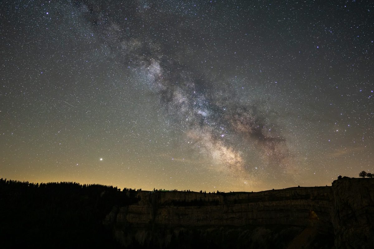 Mliečna cesta osvetľuje nočnú oblohu nad tmavým lesnatým horizontom, jasné hviezdy sa trblietajú v diaľavách.
