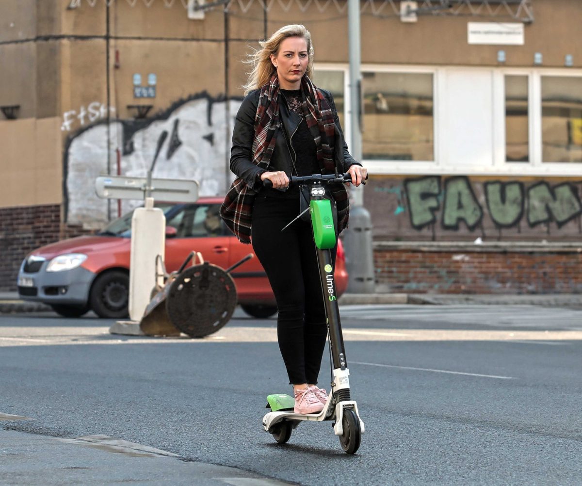 Žena na elektrickej kolobežke prechádza mestskou ulicou s grafitmi na pozadí, oblečená do šálu a bundy.