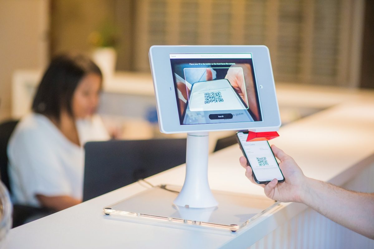 Tablet s QR kódom na obrazovke, ruka so smartfónom skenuje QR kód, žena sedí za stolom v pozadí.