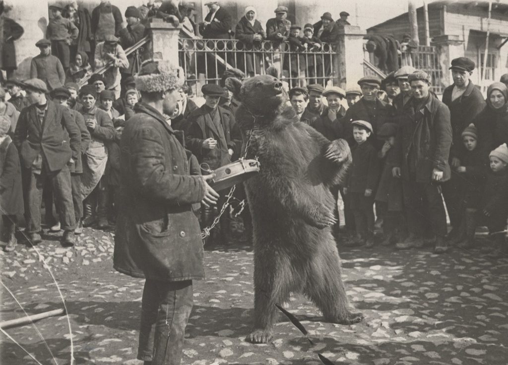 Muž s medveďom na reťazi pred davom ľudí na ulici, historická fotka ukazujúca tradičnú zábavu.