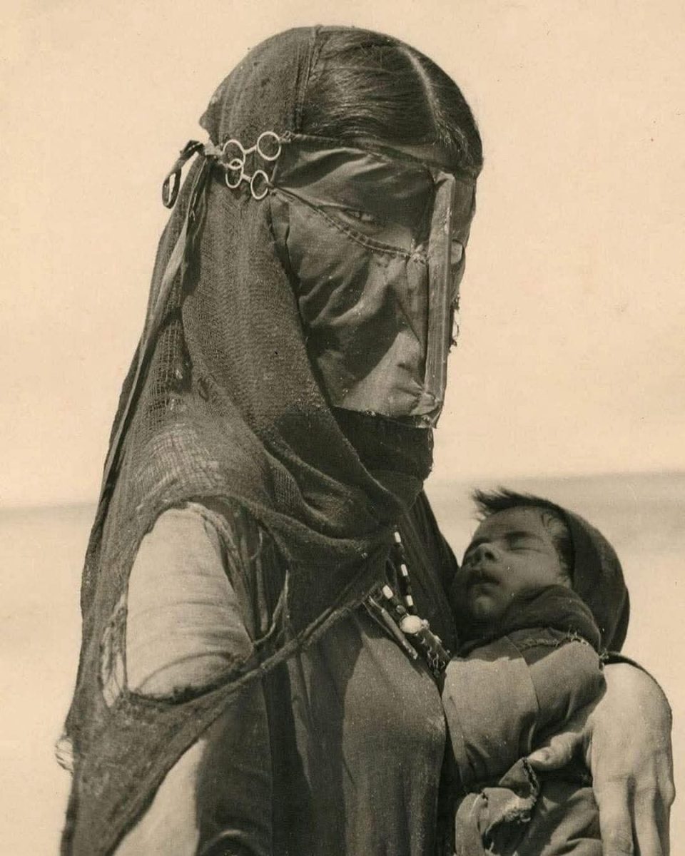 Žena v tradičnom odeve nesie spiace dieťa na púšti, historická fotografia.