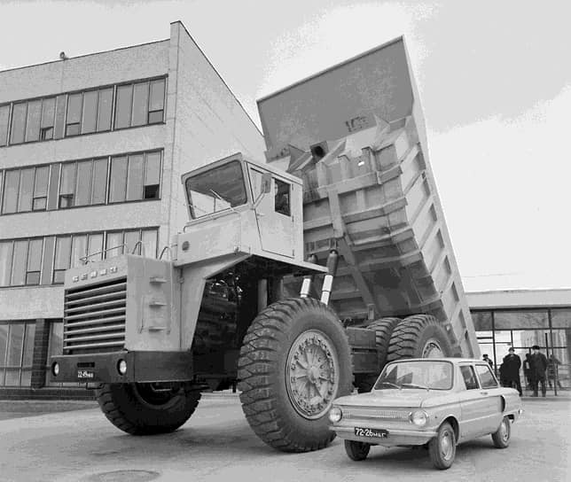 Obrovský nákladný automobil vedľa malého osobného auta pred modernou budovou.