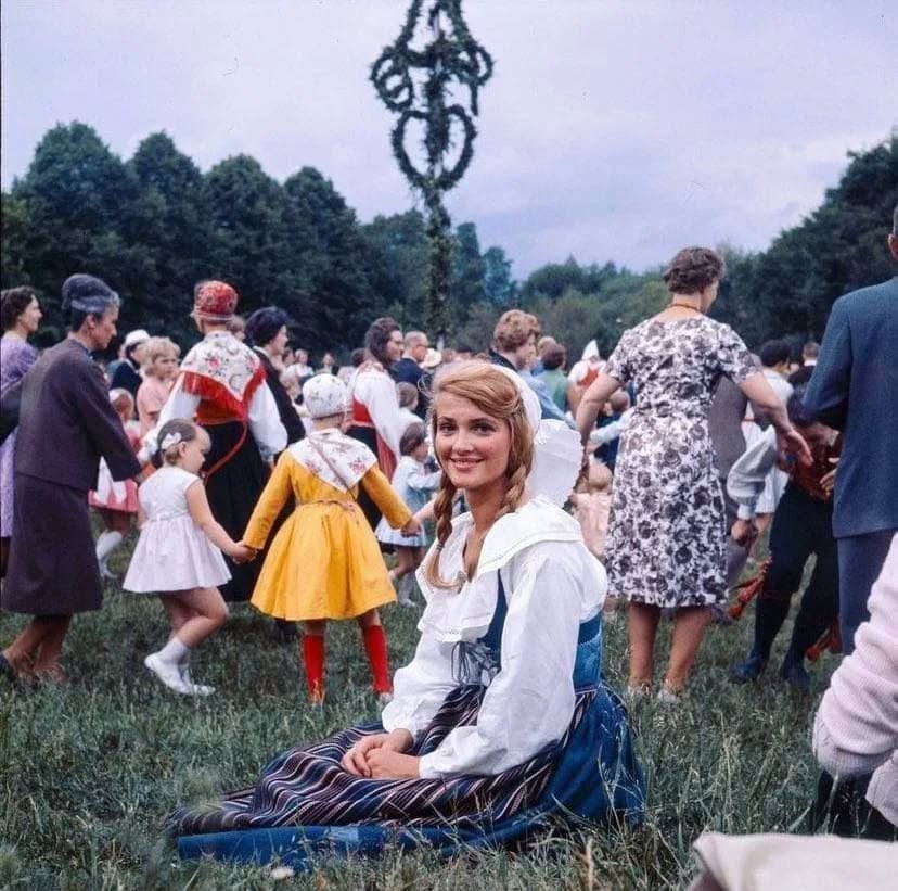 Žena v kroji sedí na lúke počas tradičného festivalu, okolo tancujú ľudia v kruhu.