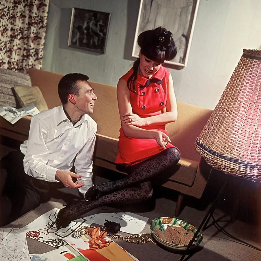 Muž a žena v retro oblečení sedí na podlahe s kresbami, usmiati pri lampáši v obývačke.