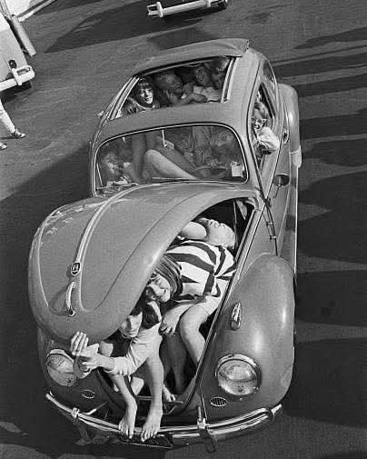 Skupina ľudí natlačená v malom aute Volkswagen Beetle na parkovisku, klasický čiernobiely záber.