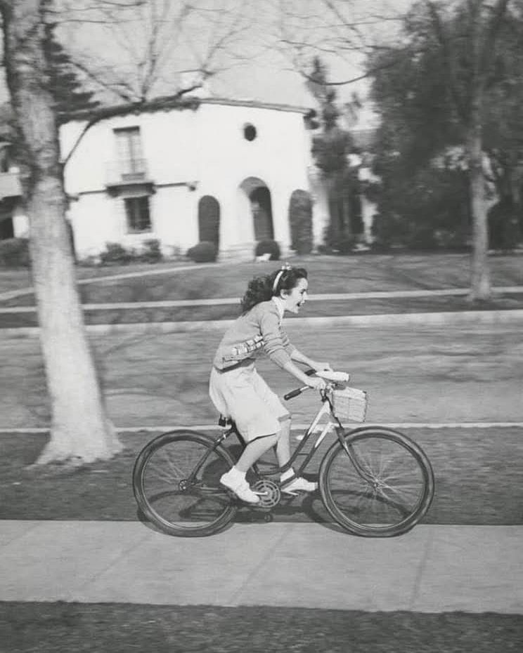 Dívka jazdící na bicykli po chodníku pred historickým domom, záber v čierno-bielej farbe.