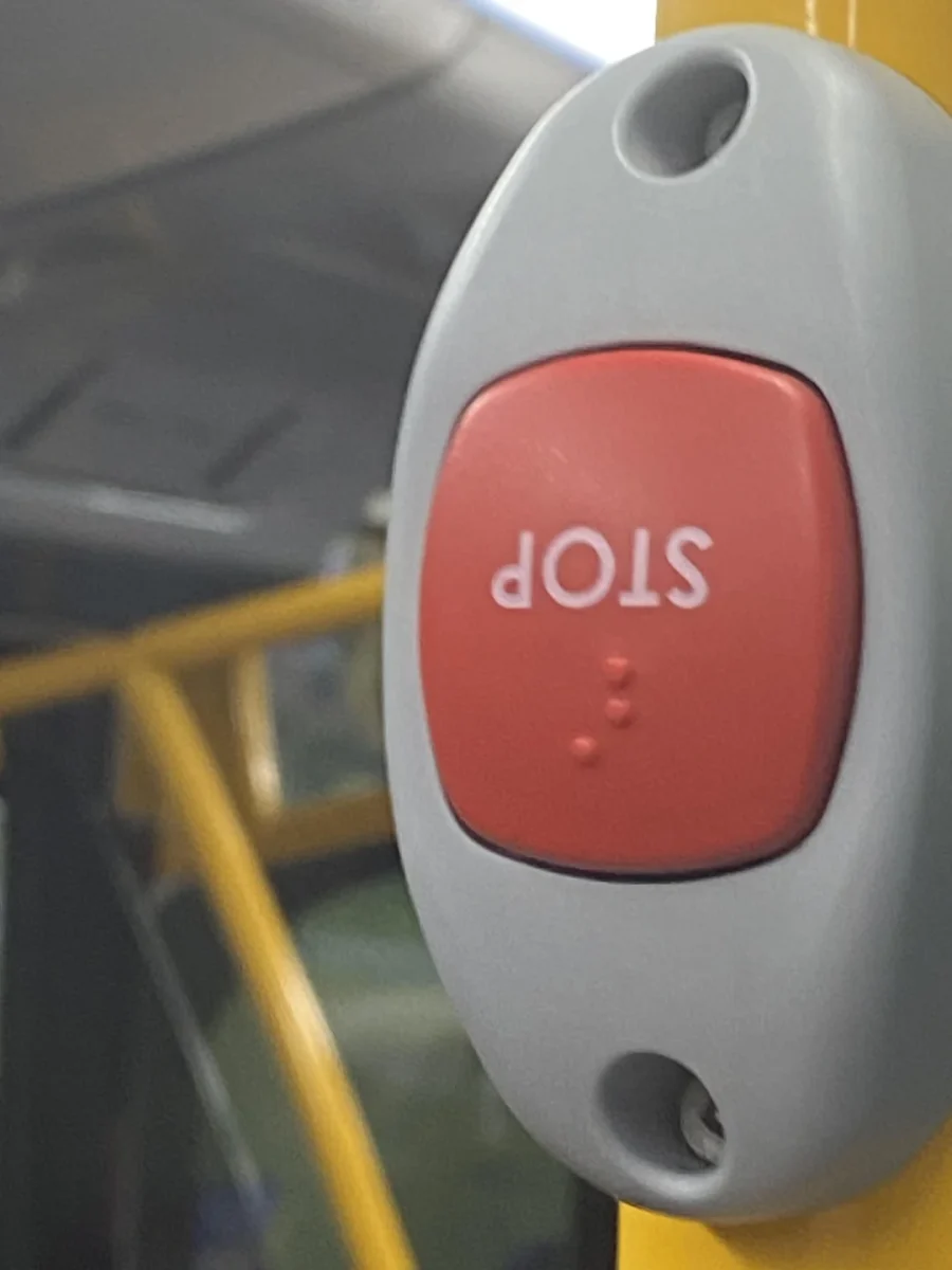 Červené tlačidlo na zastavenie autobusu so značkou STOP a Braillovým písmom na sivom podklade.