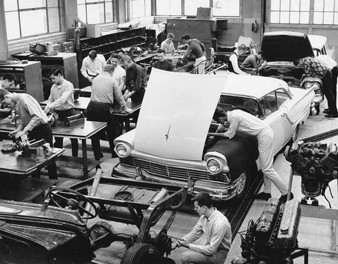 Študenti pracujúci na automobiloch v dielni počas technickej výučby, historická čiernobiela fotografia.