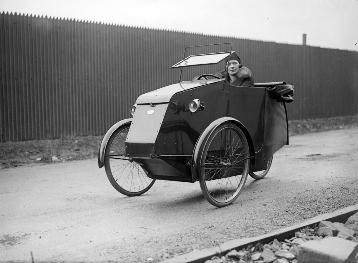 Historický trojkolesový automobil s vodičom, zachytený na ceste pred dreveným plotom.