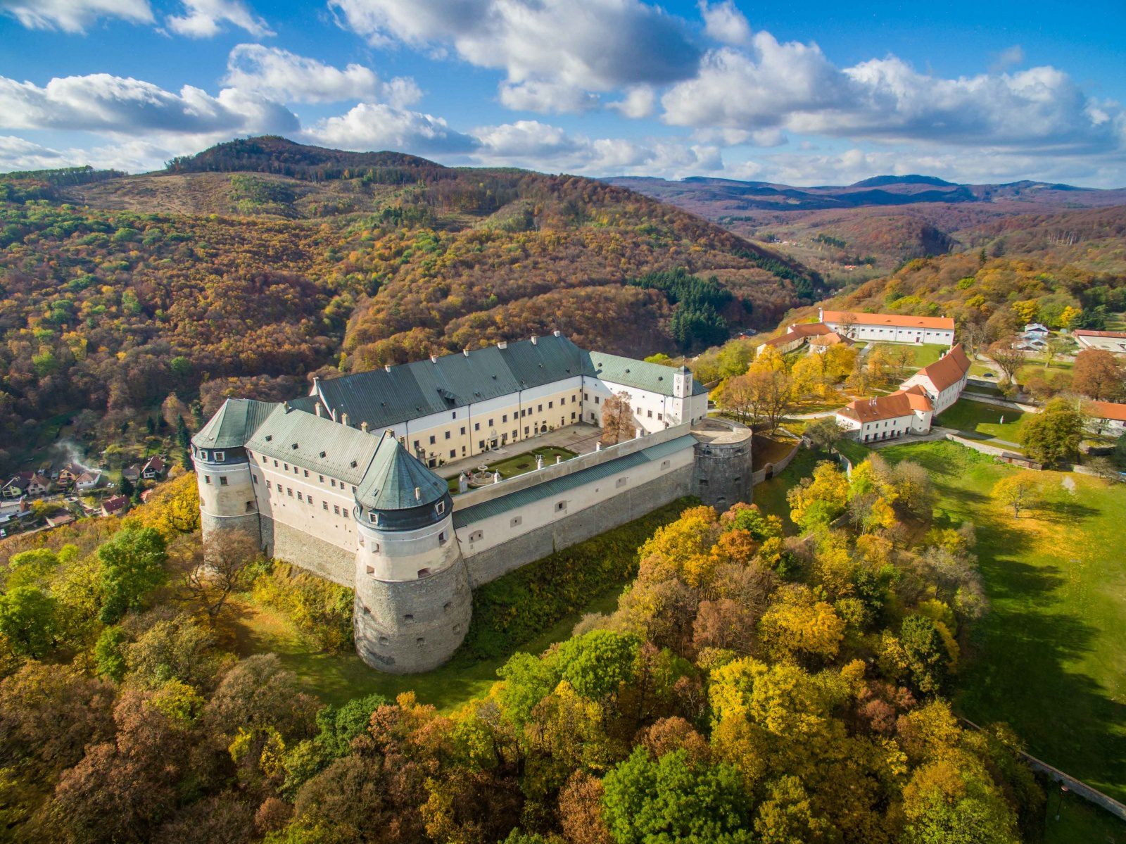 Letecký pohľad na hrad obklopený jesennou krajinou a lesmi, pod modrou oblohou s oblakmi.