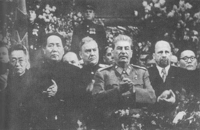 Ľudia v historických vojenských uniformách a civilnom oblečení na slávnostnom zhromaždení s kvetinovou výzdobou.