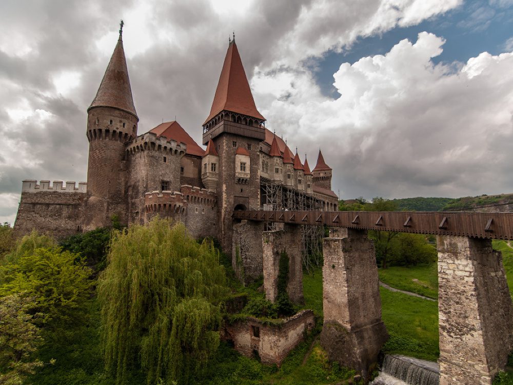 Stredoveký hrad s červenými vežami nad zelenou krajinou a kamenným mostom, dramatické oblaky na oblohe.