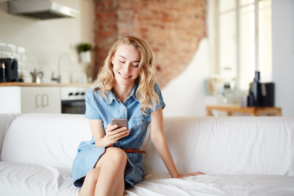 Žena v džínových šatách sedí na pohovke a s úsmevom používa mobilný telefón v moderne zariadenej kuchyni.
