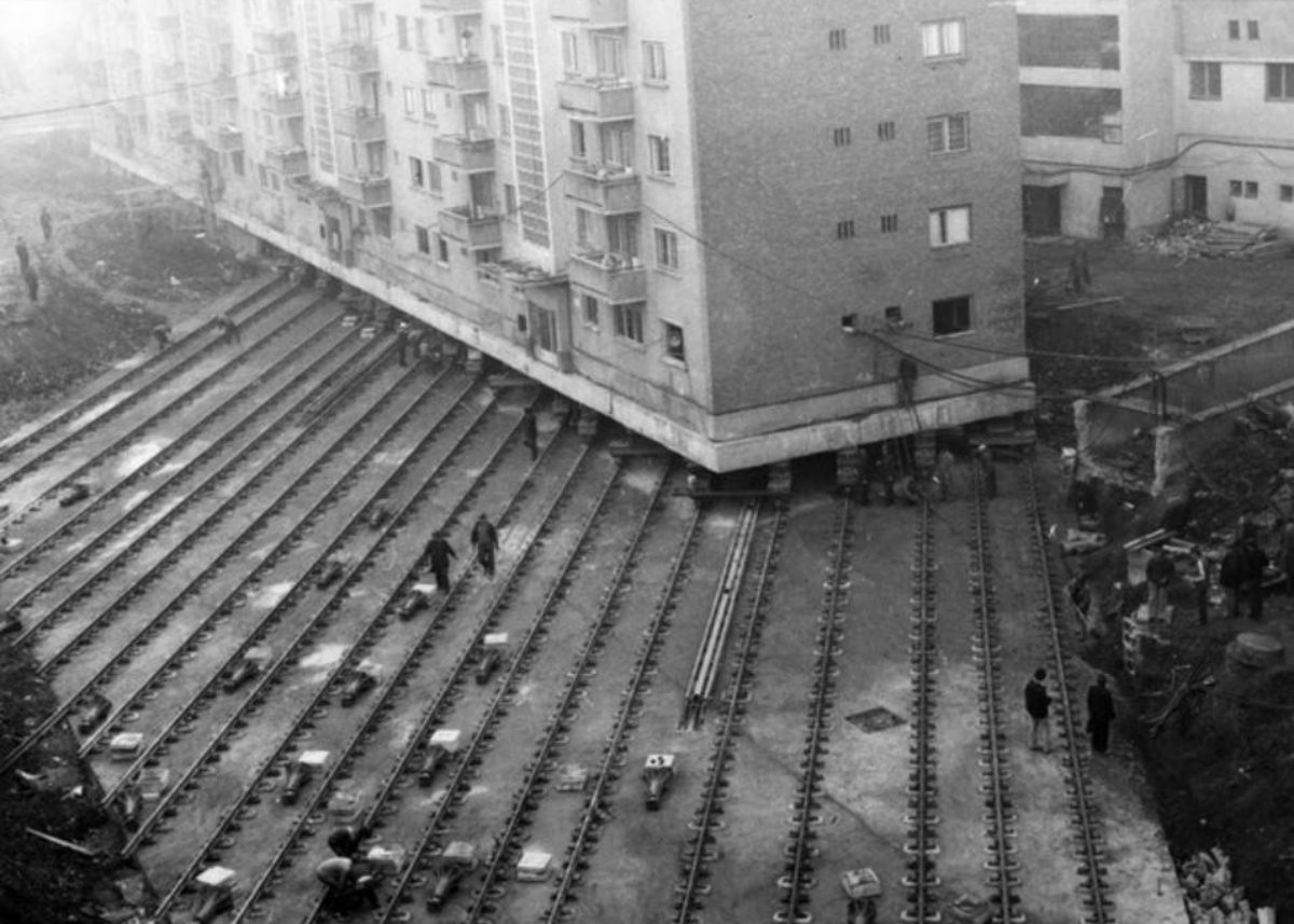 Panelák sa posúva po koľajniciach počas urbanistickej úpravy, inžinieri dohliadajú na presun budovy.