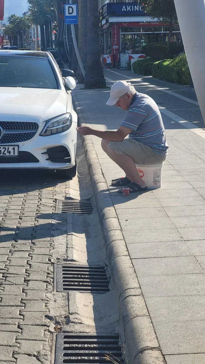 Muž sedí na vedre pri bielom aute a opravuje obrubník na slnečnej ulici.