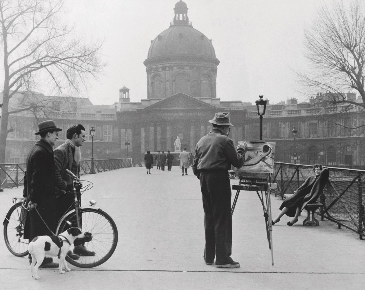 Muž maľuje na moste, kým sa ľudia prechádzajú. Vedľa stojí pán s bicyklom a psom. V pozadí historická budova.