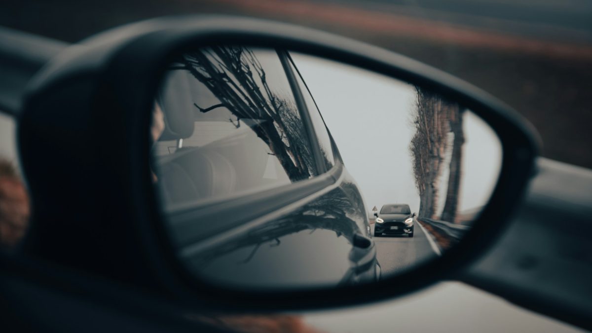 Auto v spätnom zrkadle na ceste, zahmlené počasie, stromoradie v pozadí.