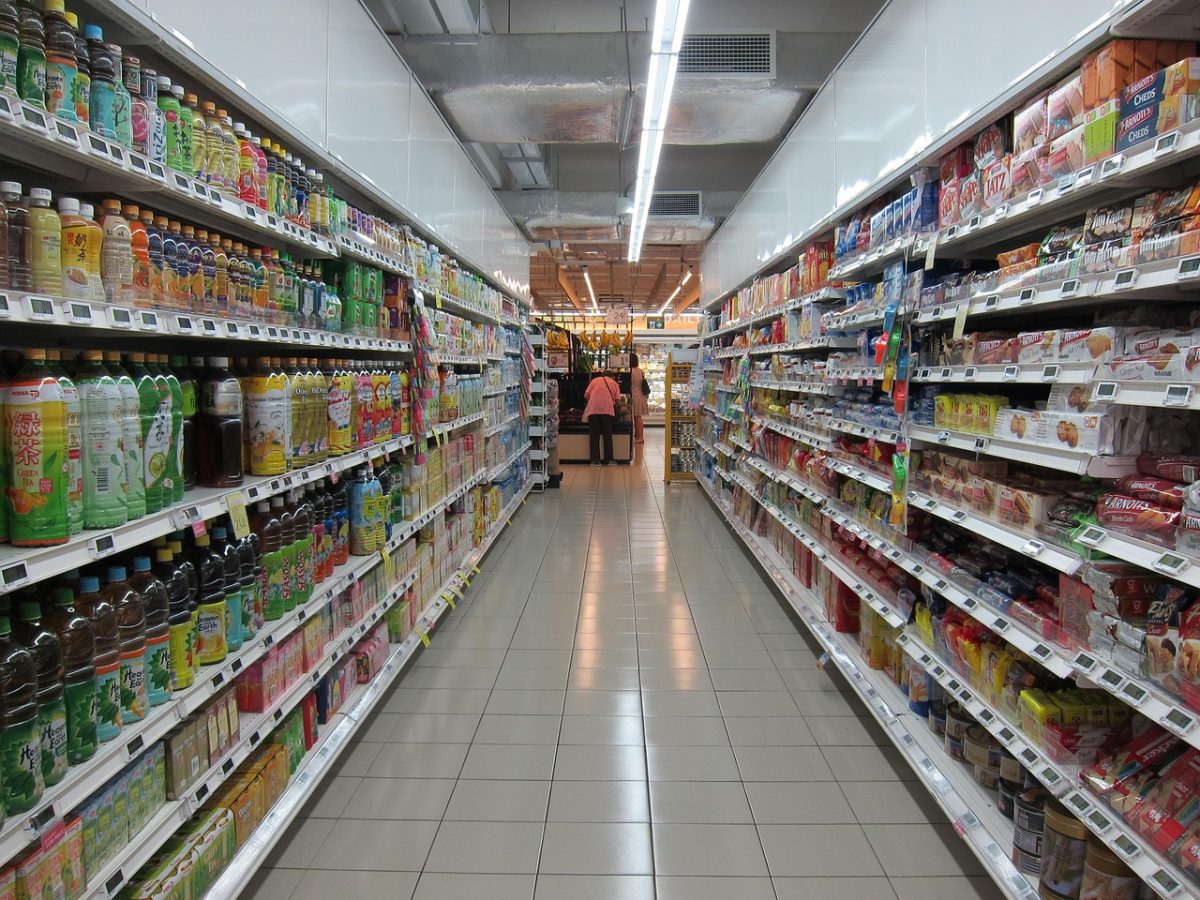 Supermarketová ulička plná nápojov a potravín na policiach, zobrazené produkty pod svetlami.