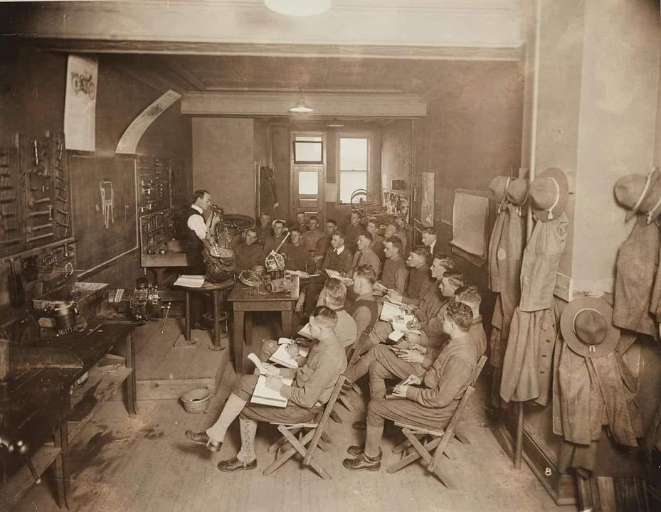 Historická trieda s mužmi na prednáške o mechanike; poznámky, nástroje a uniformy visiace na stene.
