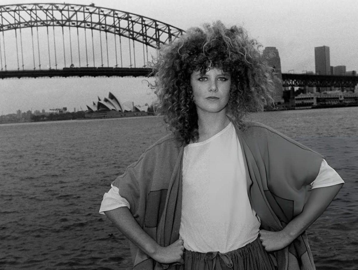 Žena s kučeravými vlasmi stojí pred Sydney Harbour Bridge a Operou, čiernobiela fotografia.