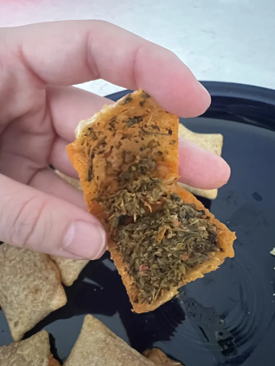 Ruka drží otvorený trojuholníkový snack s bylinkovou plnkou na tmavom tanieri.