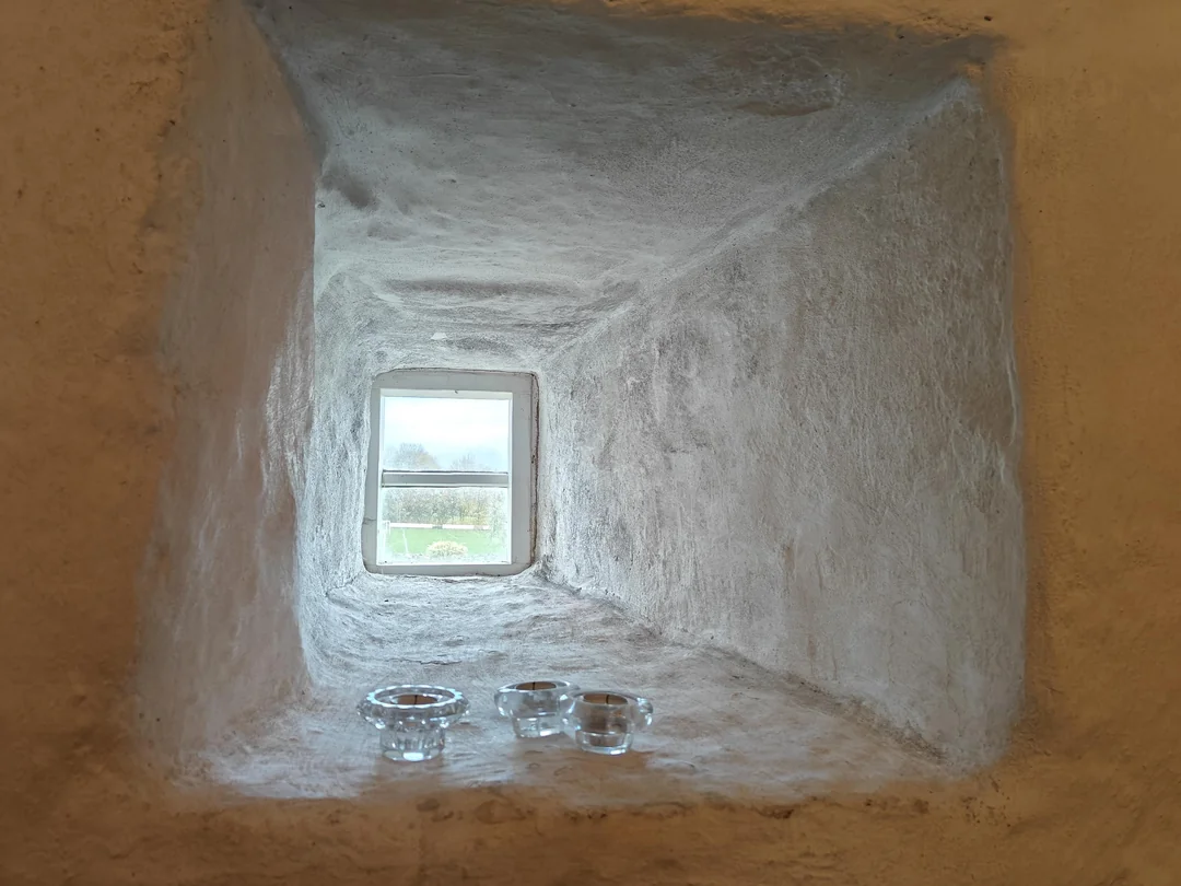Malé okno v hrubej kamennej stene s tromi sklenenými svietnikmi na parapete.