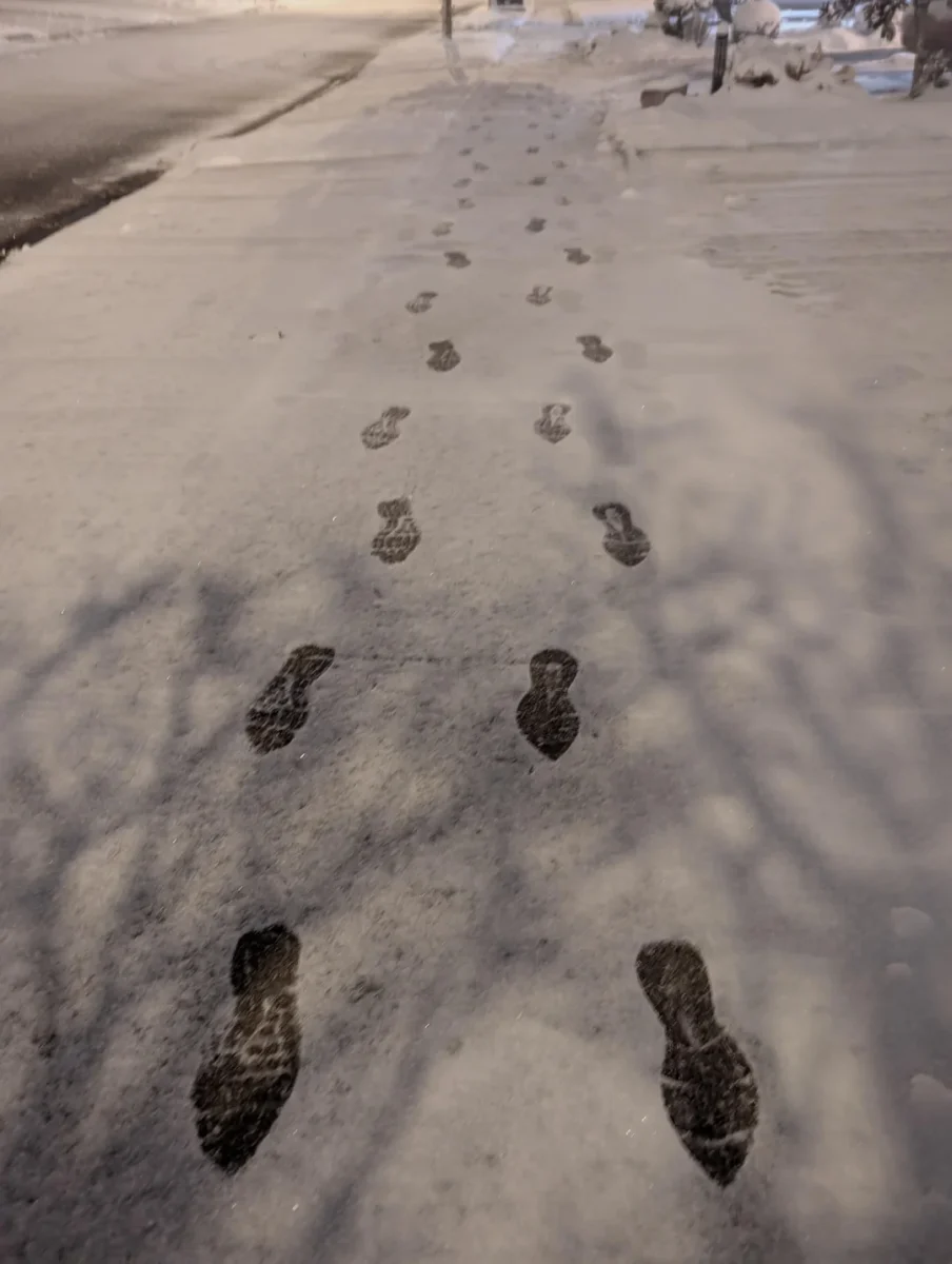 Stopy v snehu vedúce po zimnom chodníku, obklopené zasneženou krajinou a vrhajúcimi sa tieňmi stromov.