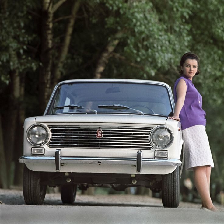 Žena opretá o biely klasický automobil na lesnej ceste.