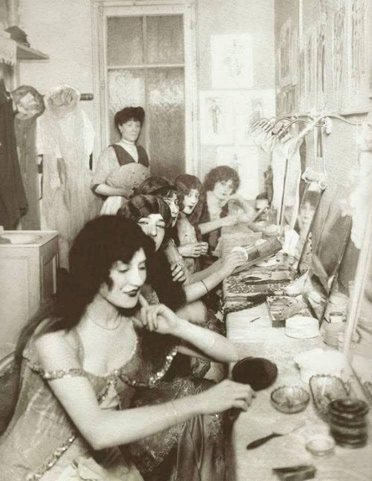 Ženy v šatni divadla aplikujú make-up pred vystúpením, historická fotografia zachytáva atmosféru zákulisia.
