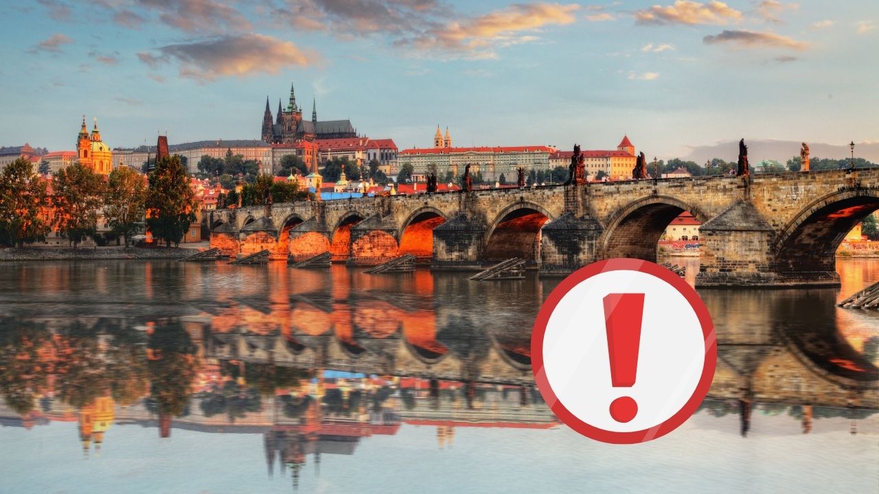 Karlov most v Prahe pri západe slnka s odrazom vo Vltave a výstražným symbolom.