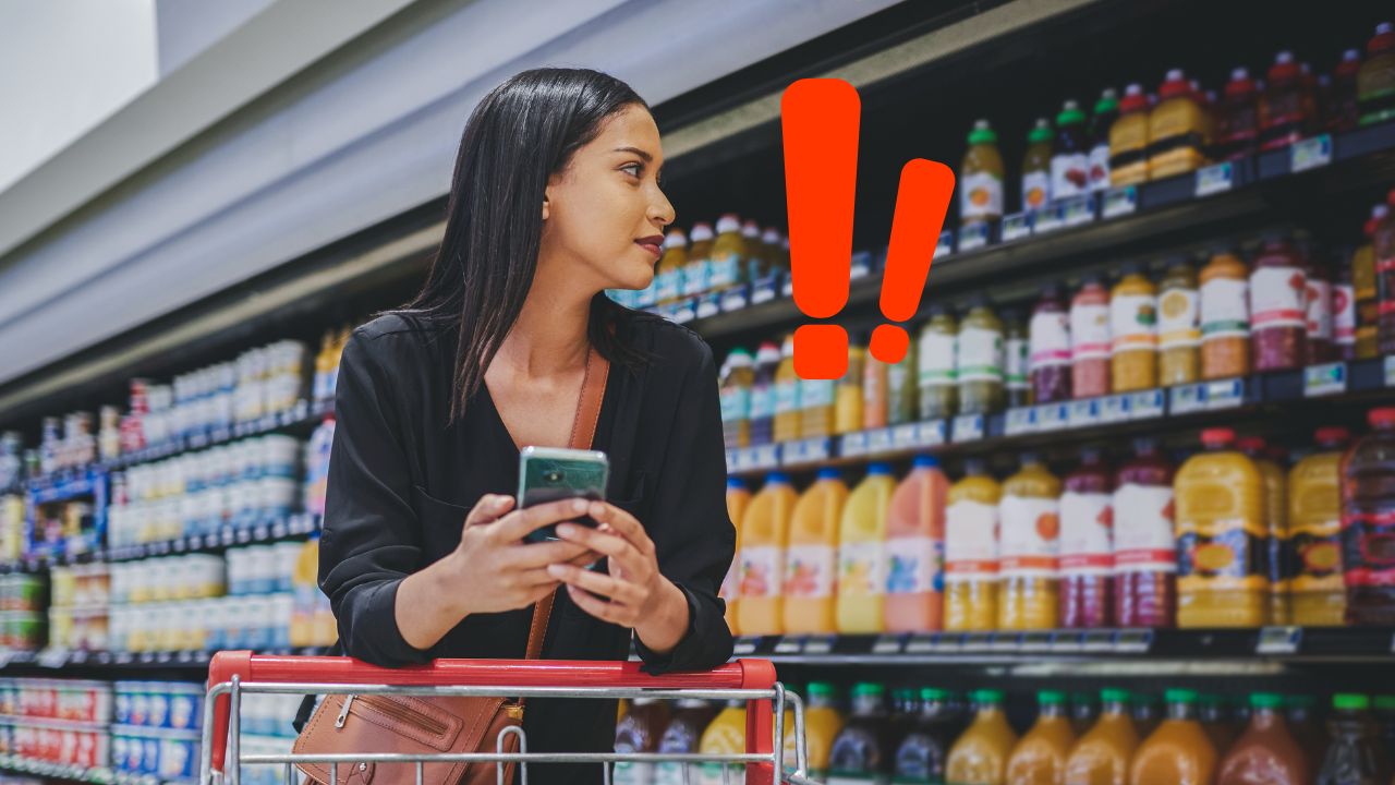 Žena s mobilom pri nákupnom košíku premýšľa o výbere džúsov v supermarkete, zameriava sa na štítky.