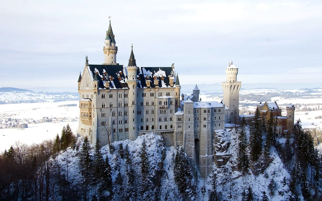 Zasnežený zámok Neuschwanstein v zimnej krajine, obklopený horami a stromami.