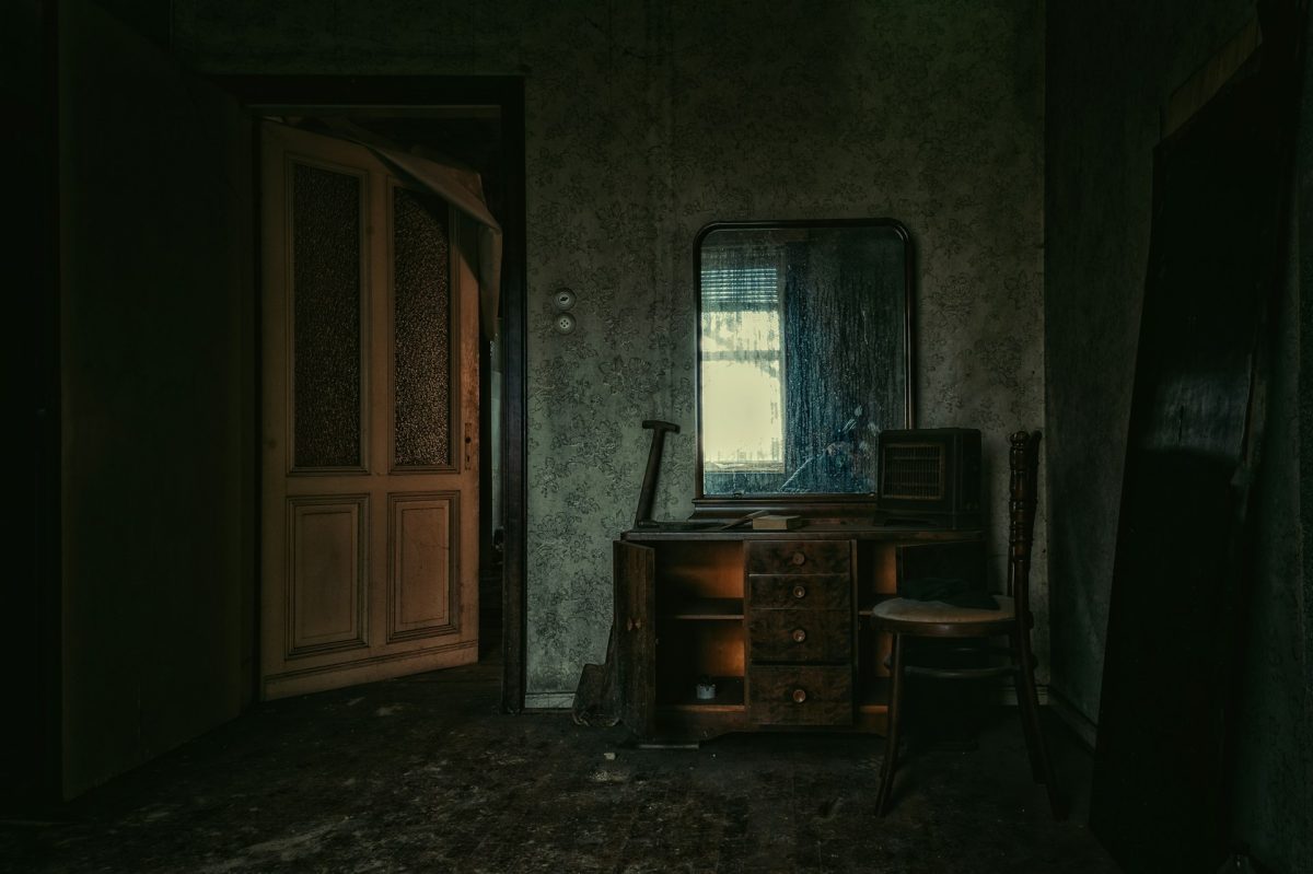 Opustená izba s dreveným nábytkom, starým zrkadlom a slabým osvetlením cez okno. Atmosféra nostalgie.