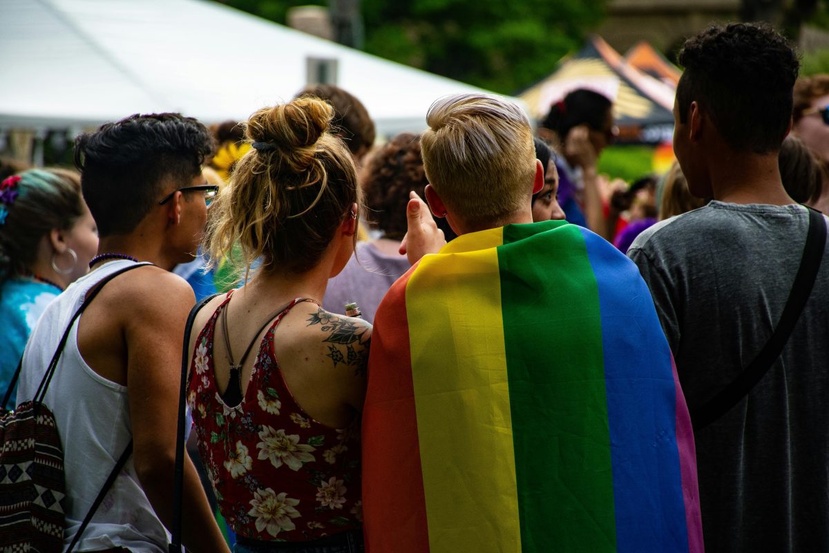 Skupina ľudí na vonkajšom podujatí, osoba v dúhovom plášti symbolizuje podporu LGBT komunity.